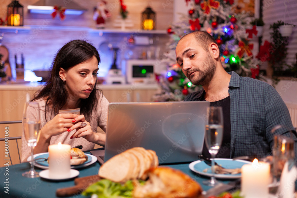 Cheerful couple shopping online xmas gift present paying with credit card on laptop computer sitting at christmas table in x-mas decorated kitchen. Happy family celebrating winter holiday