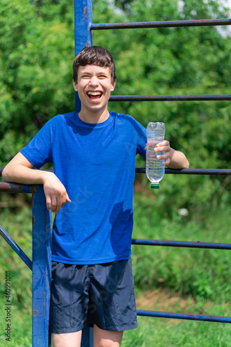 teenage boy exercising outdoors, sports ground in the yard, he stands near the horizontal bar, opens a bottle of water and drinks, healthy lifestyle