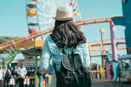 rear view of beautiful asian korean woman wearing straw hat and carrying backpack walking in front of colorful ferris wheel at amusement park on sunny day. girl looking at roller coaster outdoors. photo