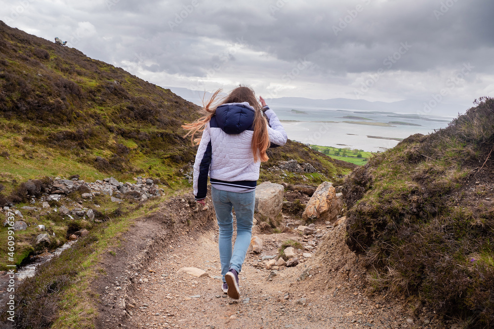 Yong teenager girl walking down a path in a mountains with beautiful scenery around her. Cloudy sky. Croagh Patrick hiking trail, Westport, county Mayo, Ireland. Outdoor activity and travel concept. Yong teenager girl walking down a path in a mountains with beautiful scenery around her. Cloudy sky. Croagh Patrick hiking trail, Westport, county Mayo, Ireland. Outdoor activity and travel concept.