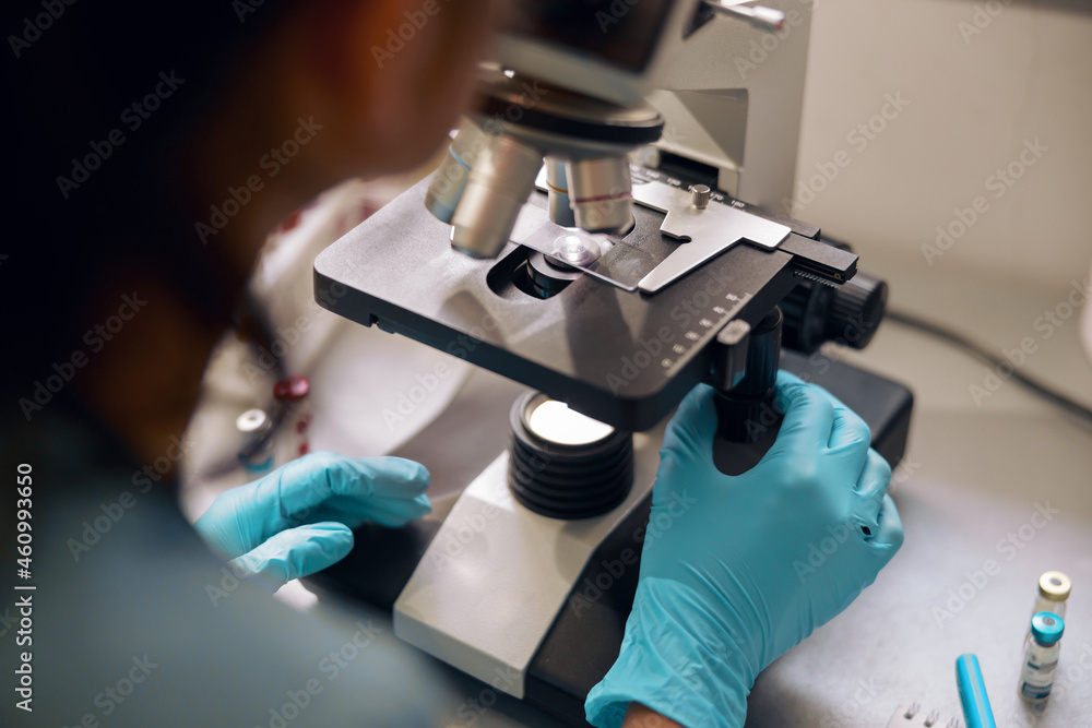 Lab assistant in latex gloves looks into microscope at sample in laboratory closeup Lab assistant in latex gloves looks into microscope at sample in laboratory closeup