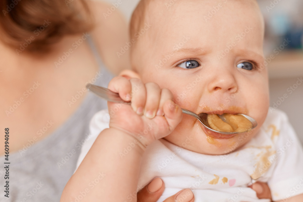 Close up portrait of cute toddler girl trying to eat fruit or vegetable puree with spoon, unknown woman holding feeding infant baby, feeding up, indoor shot. Close up portrait of cute toddler girl trying to eat fruit or vegetable puree with spoon, unknown woman holding feeding infant baby, feeding up, indoor shot.