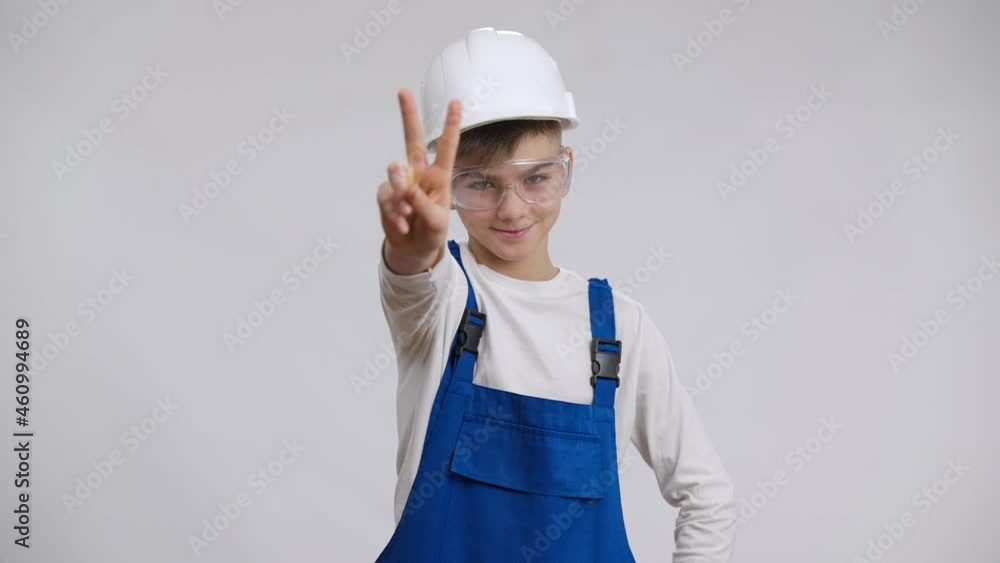 Smiling charming boy in overalls hard hat and eyeglasses showing peace gesture posing at white background. Confident Caucasian child in builder uniform looking at camera smiling. Occupation and choice Smiling charming boy in overalls hard hat and eyeglasses showing peace gesture posing at white background. Confident Caucasian child in builder uniform looking at camera smiling. Occupation and choice