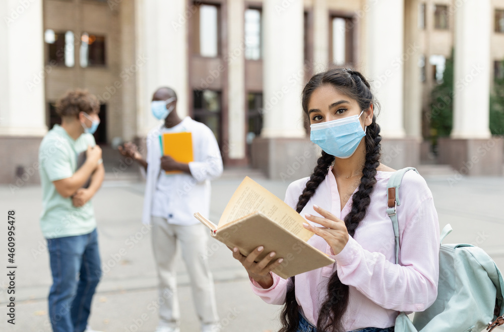 Female student wearing face mask posing with book and looking at camera, standing outdoors near university building Female student wearing face mask posing with book and looking at camera, standing outdoors near university building
