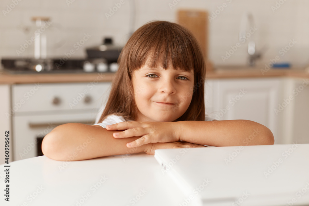 Indoor shot of charming little preschooler girl with dark hair sitting at table with folded notebook, looking at camera, posing at home in light room with kitchen set on background.