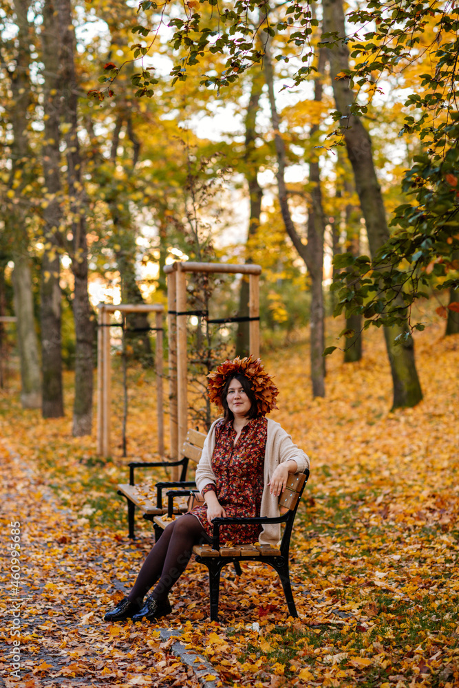 Portrait of beautiful romantic young woman with wreath of autumn yellow brown golden and red maple leaves, cute stylish girl sitting on bench in park, Selective focus, natural light, body positivity Portrait of beautiful romantic young woman with wreath of autumn yellow brown golden and red maple leaves, cute stylish girl sitting on bench in park, Selective focus, natural light, body positivity