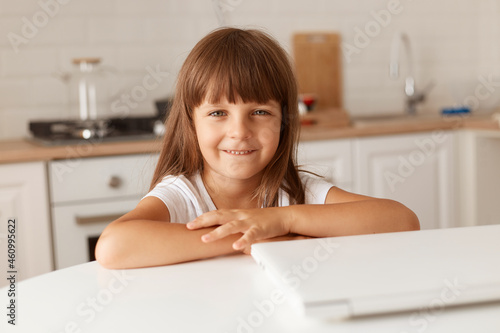 Free Download Positive little dark haired female child sitting at table near folded laptop, looking at camera with pleasant facial expression, posing at home in kitchen. - GFXLoad