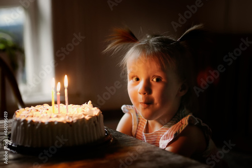 Funny baby toddler girl with two ponytails blows out three candles on the cake, dark style. Children's birthday concept