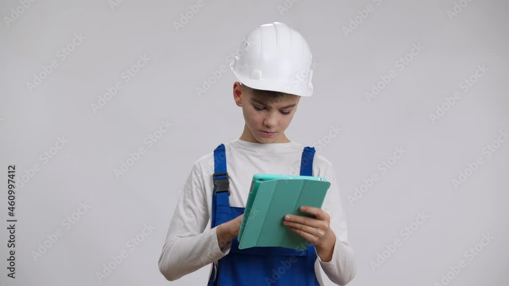 Focused serious boy in hard hat and overalls using tablet thinking and looking at camera. Portrait of confident thoughtful Caucasian child in builder uniform posing at white background. Foreman job Focused serious boy in hard hat and overalls using tablet thinking and looking at camera. Portrait of confident thoughtful Caucasian child in builder uniform posing at white background. Foreman job