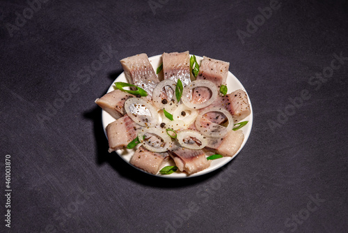 Fillet of salted herring with spices and onions on a plate. Salted sliced ​​fish on a dark background. Foods with healthy unsaturated fats, omega-3s. Top view with copy space. Healthy food
