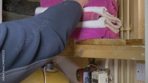 Closeup of a man's hands putting a pile of clean towels on a slatted shelf in a domestic airing cupboard shelf, above a hot water cylinder.