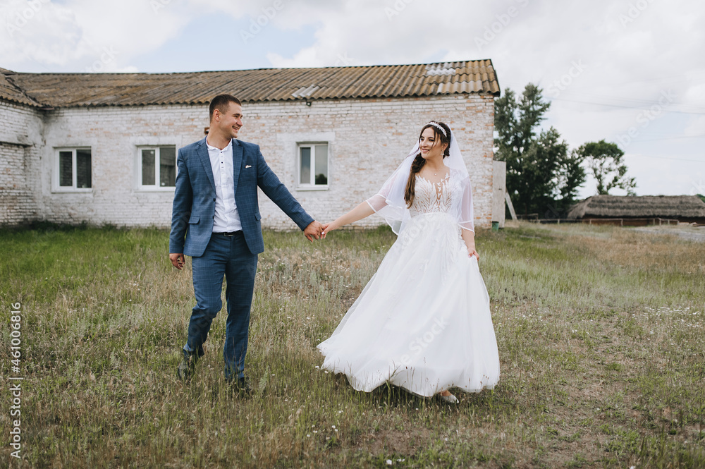 Stylish groom in a blue suit and a brunette bride in a white lace dress stroll through the village in nature on the background of a brick building, a farm. Wedding photography, portrait.