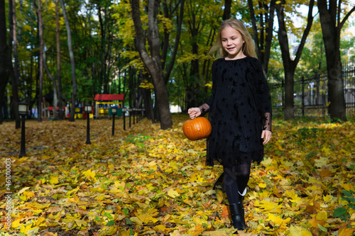 A European girl of 8 years old in a black dress with a pumpkin in her hands. Halloween in the autumn park.
