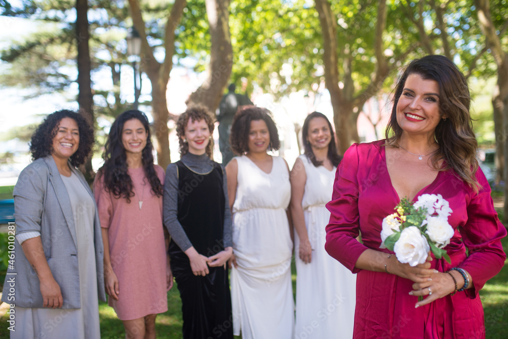 Smiling mid adult woman holding wedding bouquet. Dark haired woman in crimson dress with flowers. Women of different nationalities in festive dresses in background. Wedding, LGBT, celebration concept