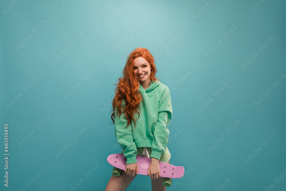 Young red-haired girl with embarrassed look smiles at camera on blue background. She has long wavy hair and light-colored clothing. In hands of fashionable lilac skateboard. Concept active recreation. Young red-haired girl with embarrassed look smiles at camera on blue background. She has long wavy hair and light-colored clothing. In hands of fashionable lilac skateboard. Concept active recreation.