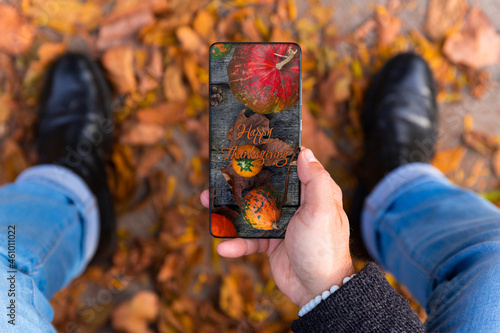 Fototapeta Naklejka Na Ścianę i Meble -  Man holding smartphone with Happy Thanksgiving photo message on the screen outdoors in a park full of autumn leaves on the ground