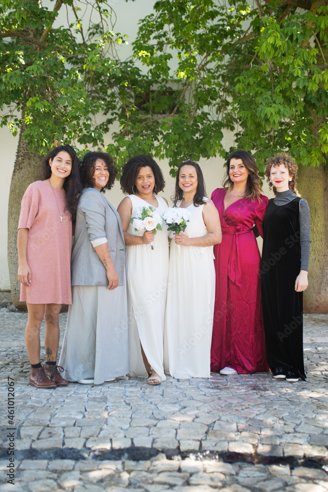 Portrait of beaming brides and guests at wedding. Women of different nationalities in festive dresses standing together and looking at camera. Wedding, LGBT, celebration concept