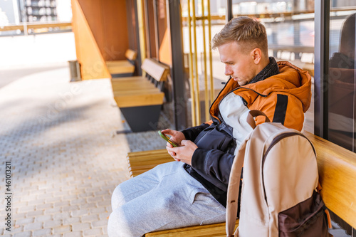 A young man uses a smartphone while sitting at a bus stop. A business man is waiting for a bus at a modern wooden bus stop