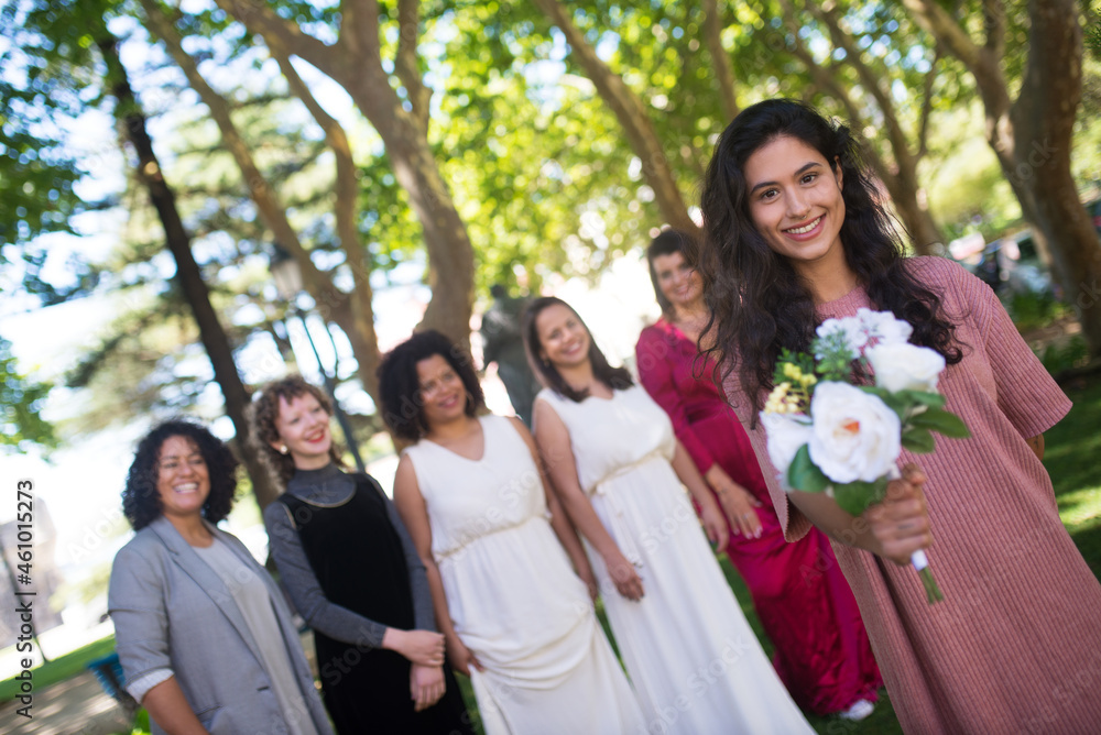 Smiling young woman holding wedding bouquet. Dark haired woman in pink dress with flowers. Women of different nationalities in festive dresses standing in background. LGBT wedding celebration concept