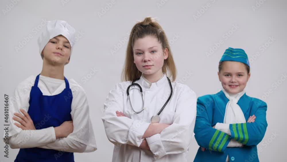 Caucasian girl in white doctor gown crossing hands looking at camera with children in chef cook and stewardess uniform posing at background. Portrait of confident little nurse choosing occupation Caucasian girl in white doctor gown crossing hands looking at camera with children in chef cook and stewardess uniform posing at background. Portrait of confident little nurse choosing occupation