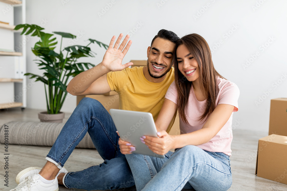 Young multinational couple chatting with friends or family online on tablet pc in new apartment. Relocation concept Young multinational couple chatting with friends or family online on tablet pc in new apartment. Relocation concept