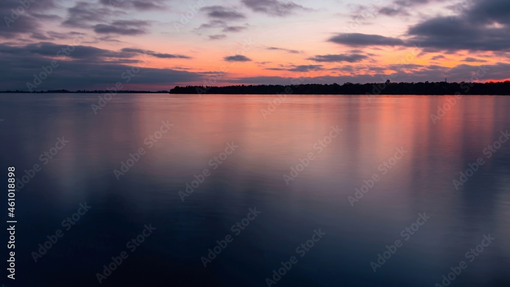 Sunset on a cloudy sky over a water surface with an island on the horizon. River landscape