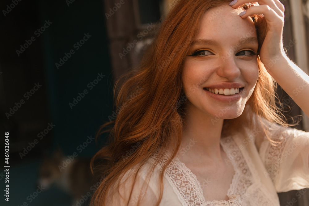 Close-up of young caucasian redhead woman with pale skin, beaming smile, dressed in white clothes. She looks away, avoiding the camera, pressing her hand to temple. Good mood concept, female beauty