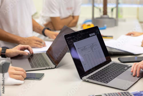 group of young university students or business people discuss, work together, note some data on notepad, laptop computer, tablet, mobile phone on desk in classroom, office or laboratory