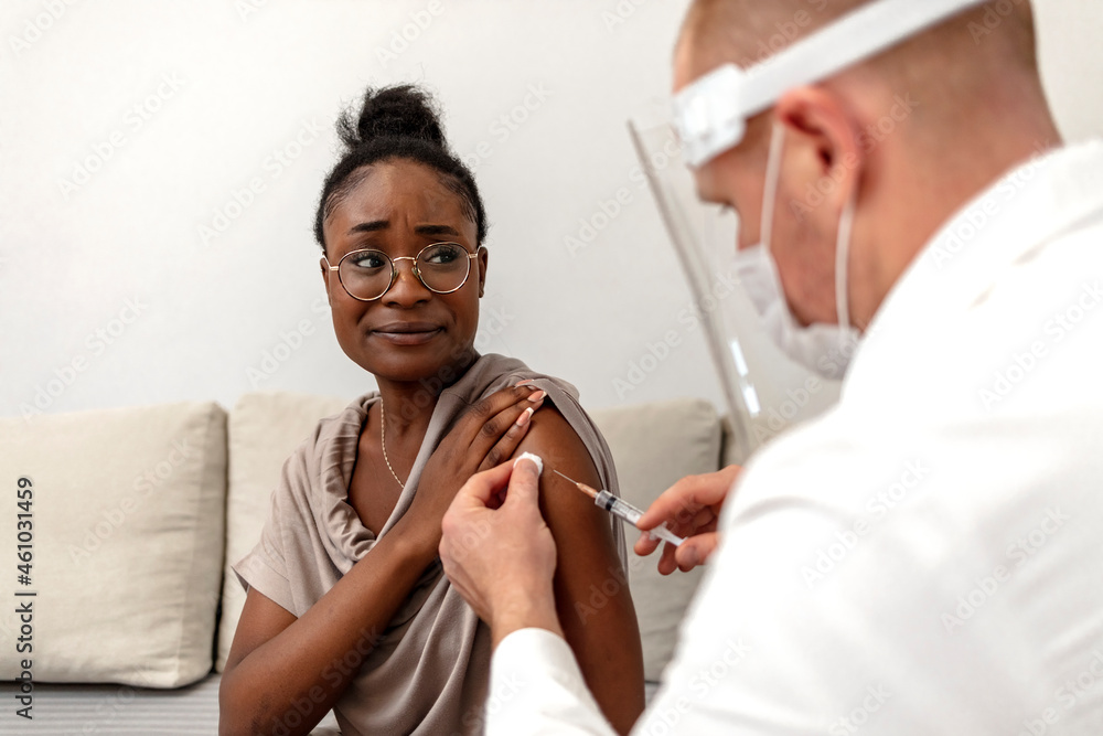 Fototapeta premium Young African woman receives a COVID-19 vaccine dose from a male healthcare worker at home. Male doctor giving a Covid-19 vaccine to an African American patient.