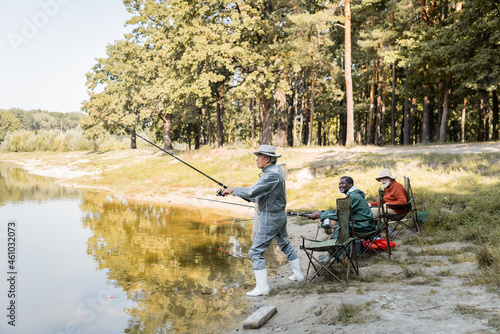 Asian man fishing near smiling multiethnic friends on lake coast