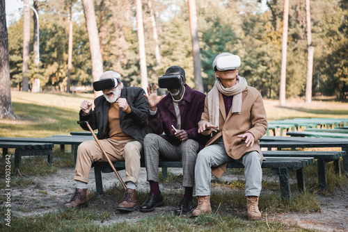 Excited interracial men in vr headsets sitting on bench in autumn park