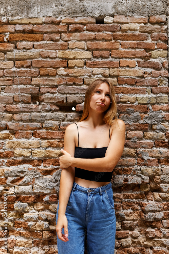 A hipster girl with long brown hair wearing a denim is looking aside while standing on a brick wall background on a street. Empty space for text o design.