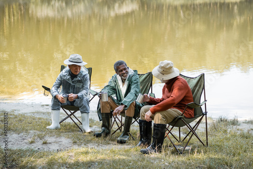 Interracial senior men in hats and fishing outfit holding thermo cups on chairs near lake