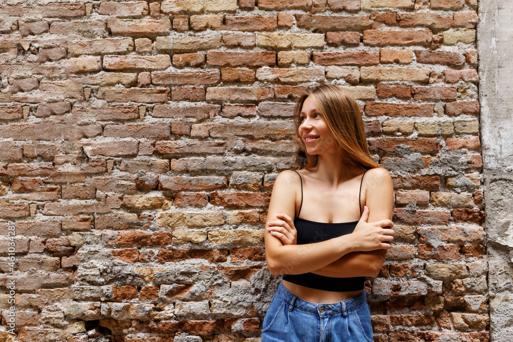 A hipster girl with long brown hair wearing a denim is looking aside while standing on a brick wall background on a street. Empty space for text o design. A hipster girl with long brown hair wearing a denim is looking aside while standing on a brick wall background on a street. Empty space for text o design.