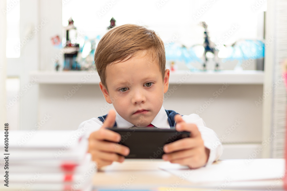 A cute first-grader boy in a school uniform on a light background at home sits at a desk with a smartphone in his hands. Selective focus. Close-up. Portrait A cute first-grader boy in a school uniform on a light background at home sits at a desk with a smartphone in his hands. Selective focus. Close-up. Portrait