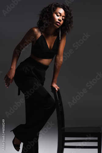 curly african american woman in trendy outfit posing on wooden chair on dark grey