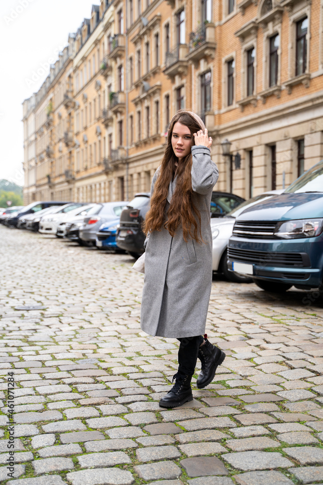 Fototapeta premium Portrait of young brunette caucasian woman dressed casually in brown hoodie and coat walking on street near residential houses and cars on cloudy early autumn day. Holiday, leisure, lifestyle