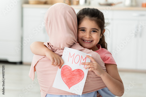 Adorable Little Girl Holding Greeting Card And Embracing Muslim Mom In Hijab