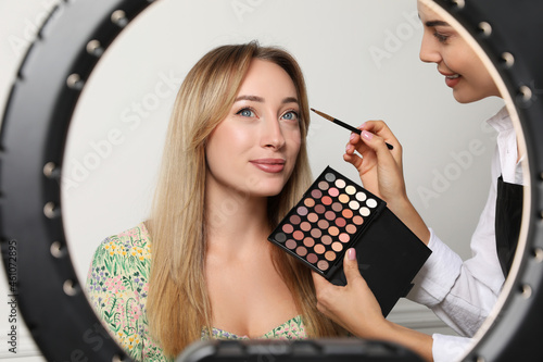 Professional makeup artist working with beautiful young woman on light background  view through ring lamp