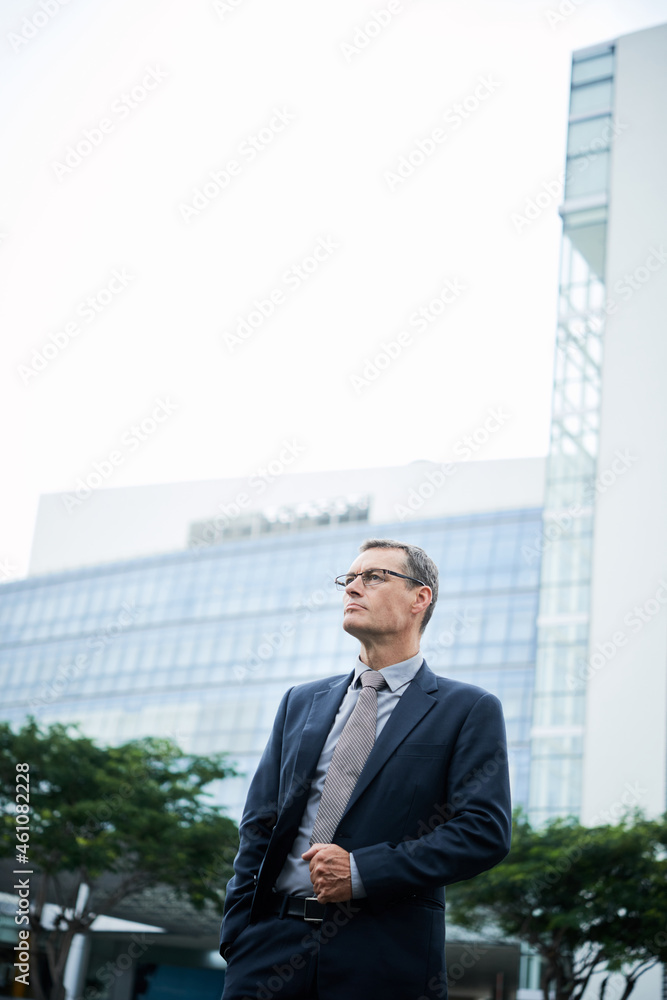 Confident mature Caucasian businessman in formal suit and glasses standing against skyscraper outdoors Confident mature Caucasian businessman in formal suit and glasses standing against skyscraper outdoors