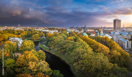 Park in Riga with trees in autumn colors. Colorful sunset over the city panorama. Downtown in background.