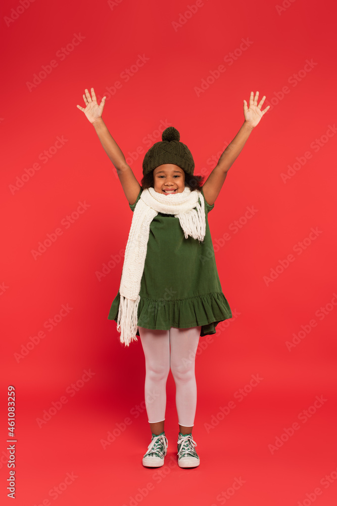 African american kid in warm hat and knitted hat waving at camera on red background