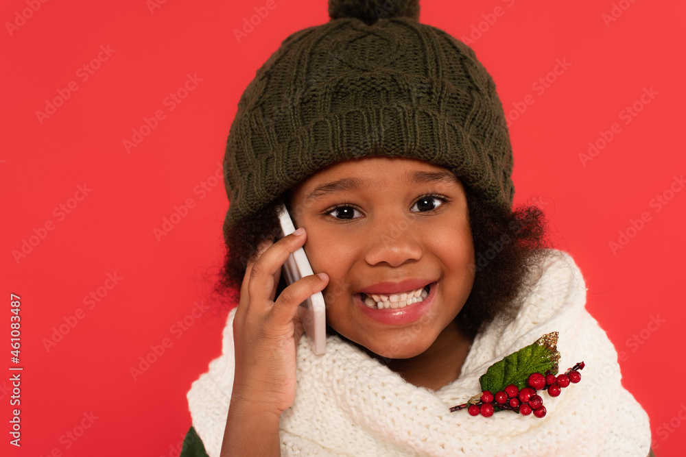 Happy african american girl in hat and scarf with mistletoe talking on mobile phone isolated on red Happy african american girl in hat and scarf with mistletoe talking on mobile phone isolated on red