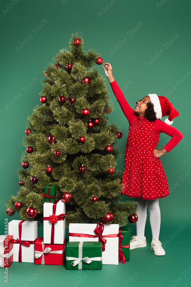 Side view of smiling african american girl in santa hat pulling hand at ball on christmas tree with gifts on green background Side view of smiling african american girl in santa hat pulling hand at ball on christmas tree with gifts on green background