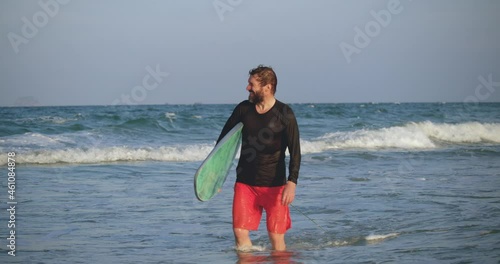 Portrait of a handsome senior sportsman at the age carrying a surfboard on the beach. A retired athlete enters the sea, goes to ride on the waves on surfing.