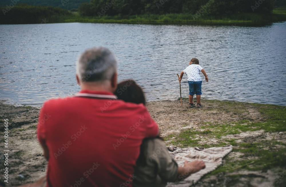 Fototapeta premium a little boy is playing by the water and his parents are watching him in the foreground on a cloudy day, rear view the average plan