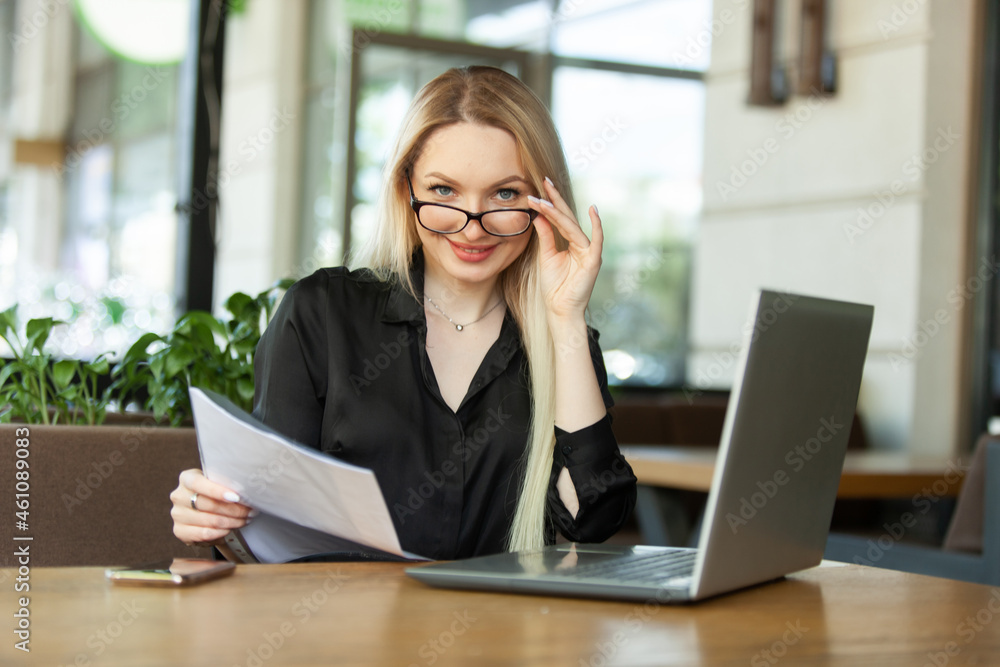 Blond Caucasian business woman examines sheets of documents and sits at a table with a laptop in an outdoor cafe. Modern business woman. Lifestyle Blond Caucasian business woman examines sheets of documents and sits at a table with a laptop in an outdoor cafe. Modern business woman. Lifestyle