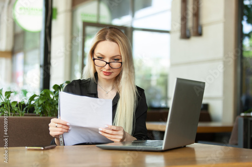 Blond Caucasian business woman examines sheets of documents and sits at a table with a laptop in an outdoor cafe. Modern business woman. Lifestyle