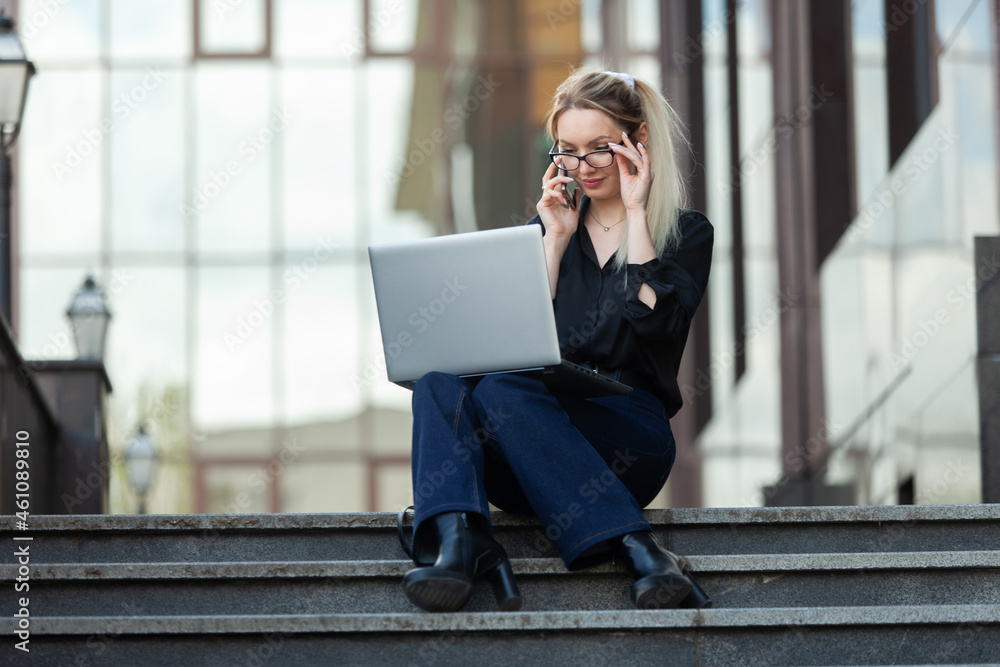 Young business woman manager talking on the phone and using a laptop while sitting on the stairs outdoors. Lifestyle. Remote work Young business woman manager talking on the phone and using a laptop while sitting on the stairs outdoors. Lifestyle. Remote work