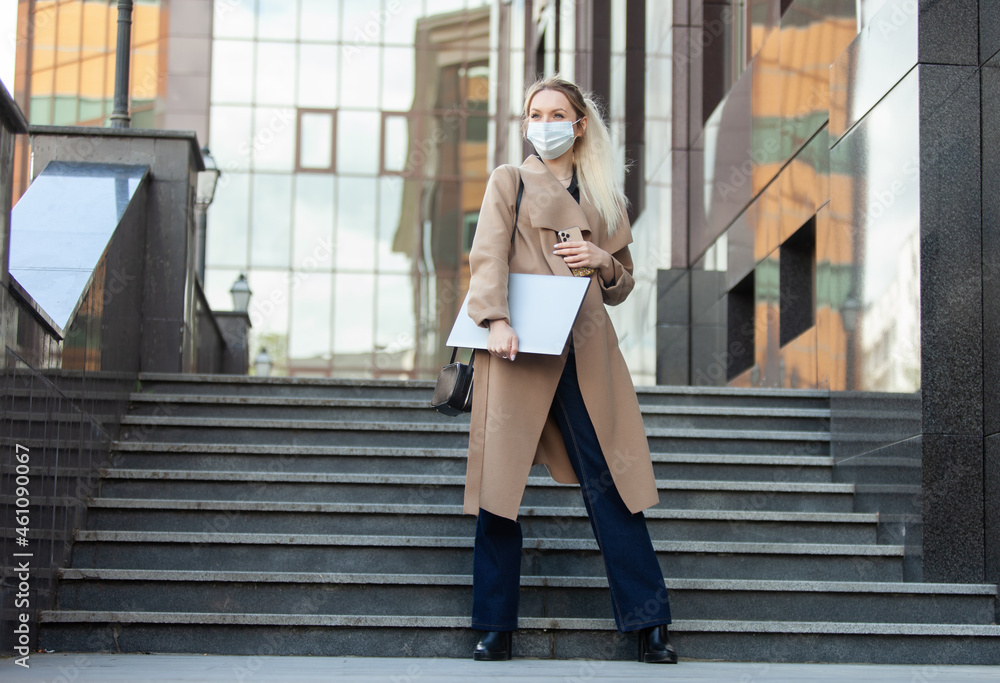 Business blonde woman in a medical mask holds a laptop in her hands on the background of the stairs Business blonde woman in a medical mask holds a laptop in her hands on the background of the stairs